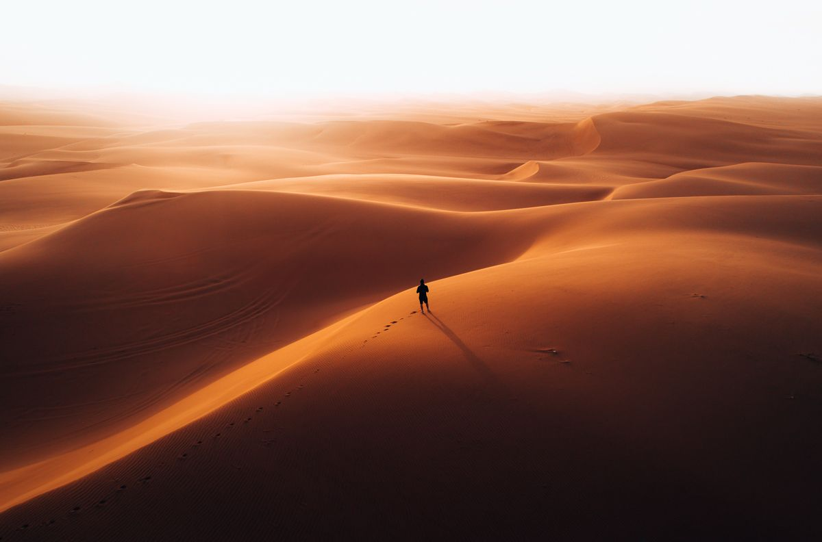 A lone figure walking across vast desert dunes at dawn — the path behind matters as much as the horizon ahead